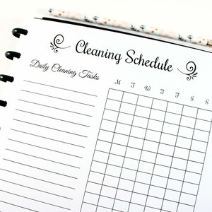 May include: A black and white Cleaning Schedule planner with a floral patterned pencil. The planner has a grid for daily tasks and the text "Cleaning Schedule" in a decorative font. The planner is open to a blank page.