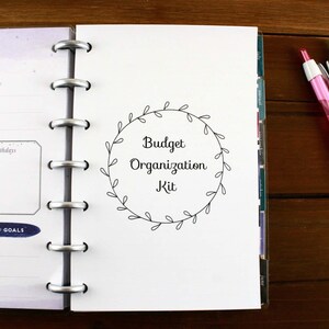 May include: Open planner with a "Budget Organization Kit" title, surrounded by a leaf wreath. The planner is open to a June page with sections for notes and goals. Two pens, one red and one green, rest on a wooden surface.