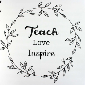 May include: A white notebook with the words "Teach Love Inspire" in black script, surrounded by a leafy wreath design. The notebook is open, revealing the spiral binding. Pencils and seashells are visible.