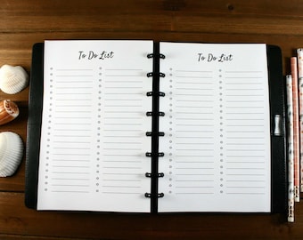 May include: An open black planner with lined pages and the words "To Do List" at the top. Three pencils with floral designs and three seashells are next to the planner on a wooden surface.