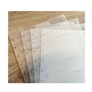 May include: A stack of clear, translucent planner dividers with a tab at the top and pre-punched holes along the side. The dividers are arranged in a fan shape, revealing a wooden surface beneath.