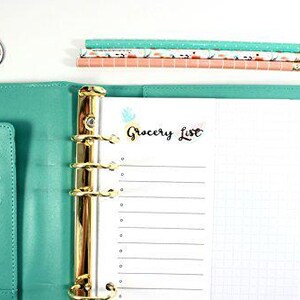 May include: A teal binder with gold accents, open to a "Grocery List" page. A floral bookmark and a sticker that says "I can & I will" are visible. Pencils with patterned designs are stacked on the top. The binder is on a white surface.