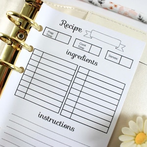 May include: A white recipe sheet with black text, held in a gold binder. The sheet includes sections for prep time, cook time, ingredients, and instructions. A floral pencil and a daisy are in the background.