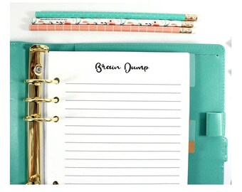 May include: A teal binder with gold hardware holds lined paper with the words "Brain Dump" printed on it. Three pencils in shades of pink and teal are stacked above the binder.