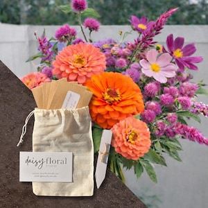 May include: A vibrant floral arrangement featuring large orange zinnias, pink cosmos, and purple blooms. A small drawstring bag and seed packets are in the foreground, with a business card that reads "daisy floral studio".