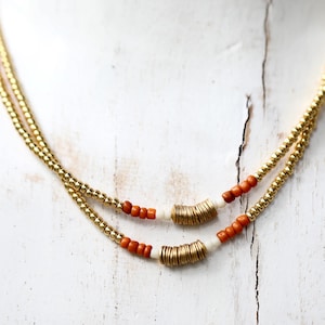 May include: A layered gold-tone necklace with small, round beads and accents of orange and white beads. The necklace features a delicate chain design and is displayed on a white surface. The necklace is a piece of jewelry.
