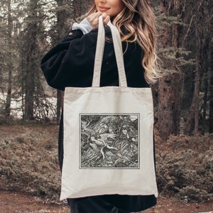 May include: A white canvas tote bag with black handles. The bag features a black and white illustration of a scene from a fantasy novel. The illustration depicts a group of fairies flying through a forest.