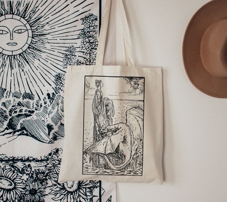 Puede incluir: Una bolsa de lona blanca con un dibujo en blanco y negro de una mujer de pie sobre una roca con un drag&oacute;n. La mujer lleva un vestido largo y tiene el pelo recogido hacia atr&aacute;s. El drag&oacute;n est&aacute; enroscado en la roca y tiene las alas extendidas.