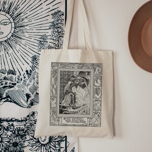 May include: A white tote bag with black and white illustration of a knight fighting a dragon. The illustration is framed with a black border and includes the text "The Dragon's Oath: When the Sword is Unsheathed, the Dragon's Fury is Unleashed."