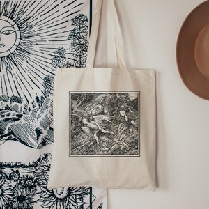 May include: A white canvas tote bag with black handles. The bag features a black and white illustration of a winged figure flying through a forest.