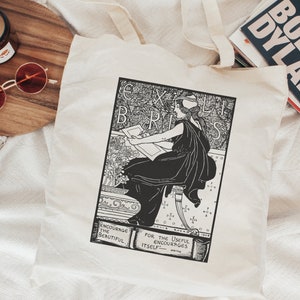 Pode incluir: Uma bolsa de tecido branca com uma ilustração em preto e branco de uma mulher sentada e desenhando. A ilustração é cercada por uma borda preta com o texto "Ex Libris" em uma fonte decorativa. Os textos "Encourage the Beautiful" e "For the Useful Encourages Itself" são impressos abaixo da ilustração.