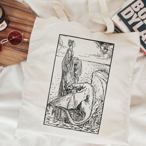 Puede incluir: Una ilustraci&oacute;n en blanco y negro de una mujer de pie sobre la espalda de un drag&oacute;n. La mujer lleva un vestido largo y tiene el pelo recogido hacia atr&aacute;s. El drag&oacute;n est&aacute; en el agua y tiene las alas extendidas. La ilustraci&oacute;n est&aacute; en una bolsa de tela blanca.