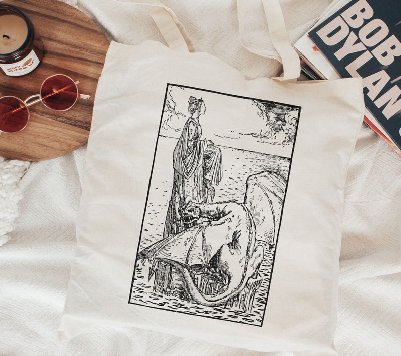 Puede incluir: Una ilustraci&oacute;n en blanco y negro de una mujer de pie sobre la espalda de un drag&oacute;n. La mujer lleva un vestido largo y tiene el pelo recogido hacia atr&aacute;s. El drag&oacute;n est&aacute; en el agua y tiene las alas extendidas. La ilustraci&oacute;n est&aacute; en una bolsa de tela blanca.