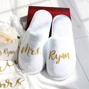 May include: White terry cloth slippers with gold glitter text reading "Mrs. Ryan". The slippers are displayed on a white surface, alongside a small white card with matching gold text. A dark red box is visible in the background.