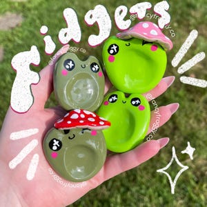 May include: Four green frog-shaped fidget toys with different mushroom-shaped lids. The lids are red and white with pink polka dots. The toys are on a hand with the text "Fidgets @fancyfroggy.co" written in white on a green background.
