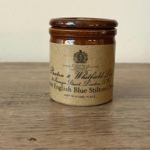 Peut inclure: Un pot cylindrique en céramique avec un couvercle vernissé brun foncé et un corps beige moucheté. L'avant présente un blason noir imprimé et le texte Paxton & Whitfield Ltd, 93 Jermyn Street, London, S.W., Best English Blue Stilton Cheese.