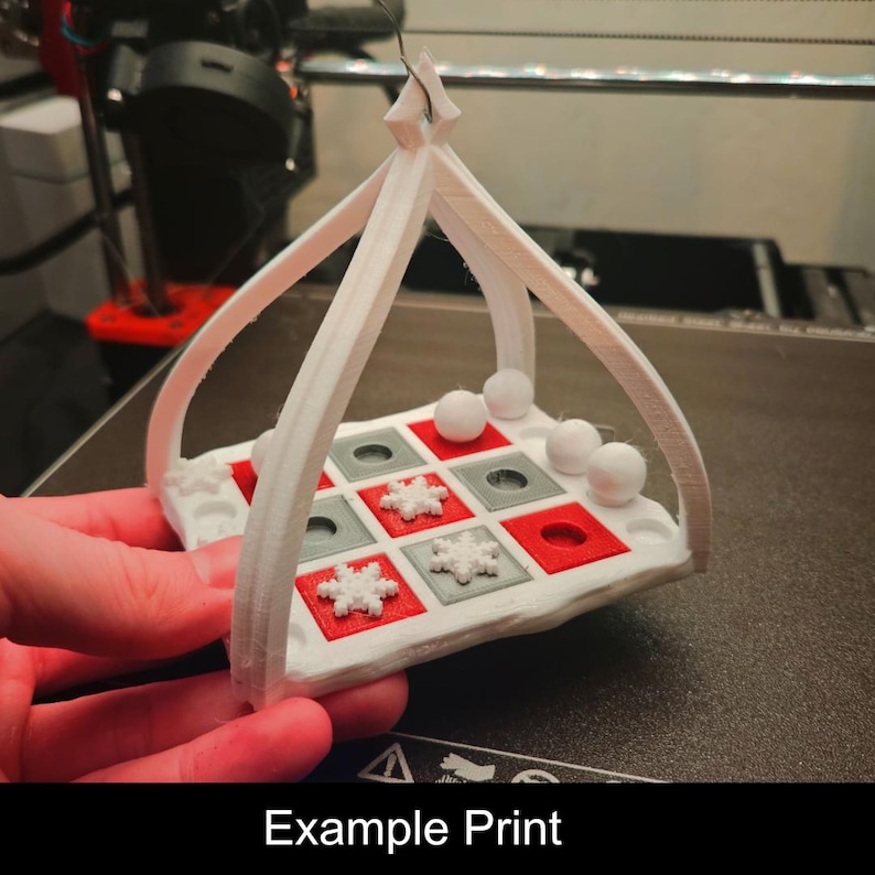 Puede incluir: Un adorno de juego de tres en raya impreso en 3D en color blanco con un marco para colgar. El tablero presenta espacios cuadrados empotrados que alternan entre rojo y gris. Incluye tres piezas con forma de copo de nieve y tres esferas.