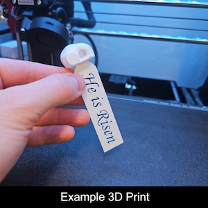 May include: A white 3D-printed object with a rounded top and two circular holes, attached to a rectangular tag. The tag reads "He is Risen" in black script. The image also includes the text "Example 3D Print".