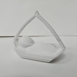 Może przedstawiać: Biała figurka wydrukowana w technologii 3D, przedstawiająca owinięte w pieluszki niemowlę leżące w małej prostokątnej kołysce. Kołyska posiada wysoki, łukowaty baldachim, kt&oacute;ry łączy się w jednym punkcie nad środkiem, tworząc tr&oacute;jkątną ramę.