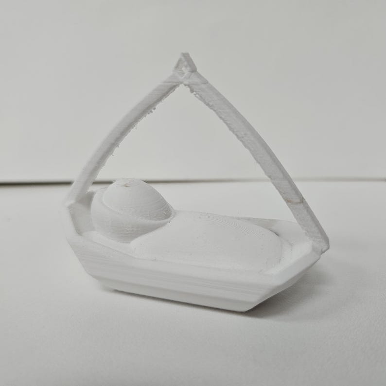 Może przedstawiać: Biała figurka wydrukowana w technologii 3D, przedstawiająca owinięte w pieluszki niemowlę leżące w małej prostokątnej kołysce. Kołyska posiada wysoki, łukowaty baldachim, kt&oacute;ry łączy się w jednym punkcie nad środkiem, tworząc tr&oacute;jkątną ramę.