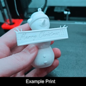 Puede incluir: Un adorno de mu&ntilde;eco de nieve impreso en 3D de color blanco, con un sombrero y un letrero rectangular horizontal sostenido al frente. El letrero tiene el texto en relieve Merry Christmas!!! con tres signos de exclamaci&oacute;n.