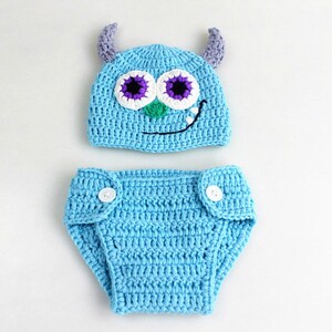 May include: A blue crocheted baby hat and diaper set with a monster face design. The hat has purple horns, white eyes, and a green mouth. The diaper has two white buttons.