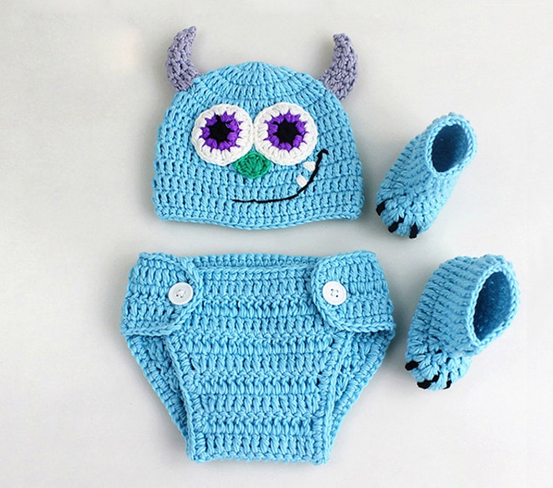 May include: A blue crocheted baby hat, diaper cover, and booties set with monster face details. The hat has purple horns and big eyes with white and purple accents. The booties have a claw-like design.