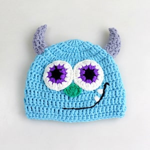 May include: A blue crocheted hat with purple and white eyes and a green nose. The hat has two purple horns on top.