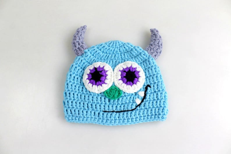 May include: A blue crocheted hat with purple and white eyes and a green nose. The hat has two purple horns on top.