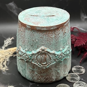 May include: A teal and copper colored ceramic piggy bank with an ornate design and a slot on the top. The piggy bank is sitting on a black surface with several US quarters scattered around it.