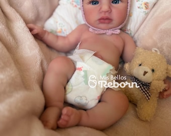 May include: A reborn baby doll with blue eyes and a pink bonnet, wearing a diaper, and lying on a pink blanket. A small teddy bear with a bow tie is next to the doll. The doll's name is "Mis Bellos Reborn".