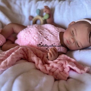 Puede incluir: Un beb&eacute; reborn durmiendo con un mono de punto rosa y una diadema rosa. El beb&eacute; est&aacute; acostado en una manta blanca con una manta rosa.