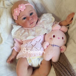 Può includere: Una bambola reborn realistica con occhi azzurri, che indossa un top in maglia rosa con colletto in pizzo bianco, una fascia con fiori rosa e un pannolino. La bambola tiene in mano un piccolo coniglietto di peluche rosa. Il testo Mis Bellos Reborn è visibile.