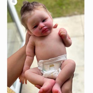 May include: A realistic reborn baby doll with light skin, rosy cheeks, and soft brown hair. The doll has detailed facial features, including dark eyes and a small mouth, and is wearing a white disposable diaper with a yellow and white graphic print.