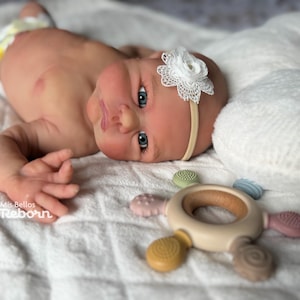 May include: A realistic reborn baby doll with blue eyes and a white lace headband. The doll is lying on a white blanket next to a wooden teething toy with colorful silicone attachments. The doll is wearing a diaper.