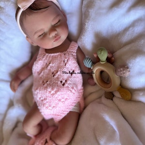 Puede incluir: Un beb&eacute; reborn durmiendo con un mono de punto rosa y una diadema rosa con lazo. El beb&eacute; est&aacute; sujetando un mordedor de madera con un dise&ntilde;o de serpiente verde. El beb&eacute; est&aacute; acostado sobre una manta blanca.