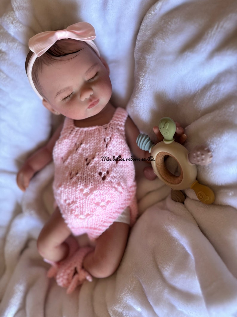Puede incluir: Un beb&eacute; reborn durmiendo con un mono de punto rosa y una diadema rosa con lazo. El beb&eacute; est&aacute; sujetando un mordedor de madera con un dise&ntilde;o de serpiente verde. El beb&eacute; est&aacute; acostado sobre una manta blanca.