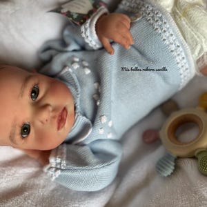 Op de afbeelding: Een realistische babypop gekleed in een lichtblauwe trui met witte randen en kleine witte bloemaccenten. De pop heeft gedetailleerde gelaatstrekken, waaronder open ogen en een licht geopende mond. Een houten en siliconen speelgoed ligt naast de pop.