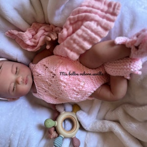 Puede incluir: Un conjunto de punto rosa con dise&ntilde;o de encaje, usado por una mu&ntilde;eca reborn dormida con una diadema rosa. La mu&ntilde;eca est&aacute; tumbada en una manta blanca con un juguete de madera.