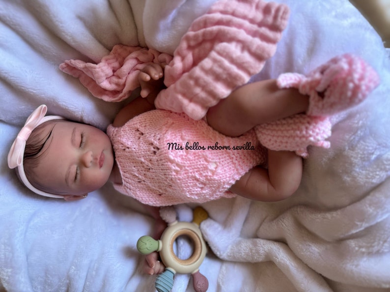Puede incluir: Un conjunto de punto rosa con dise&ntilde;o de encaje, usado por una mu&ntilde;eca reborn dormida con una diadema rosa. La mu&ntilde;eca est&aacute; tumbada en una manta blanca con un juguete de madera.