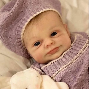 May include: A realistic baby doll with light skin and fine hair, wearing a lavender knitted beret and a matching lavender knitted sweater with cream-colored trim. The doll has dark eyes and a neutral expression, featuring a textured knit pattern.