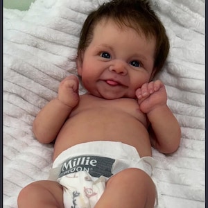 May include: A realistic reborn doll with brown hair, blue eyes, and a smiling expression with a visible tongue. The doll is wearing a white diaper with a grey waistband featuring the text Millie Moon and a small patterned print of leaves and animals.