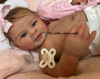 Puede incluir: Una muñeca reborn realista con ojos azules y un lazo rosa en el pelo. La muñeca está tumbada sobre una manta rosa y lleva un pañal. El texto "Mis bellos reborn sevilla" es visible.