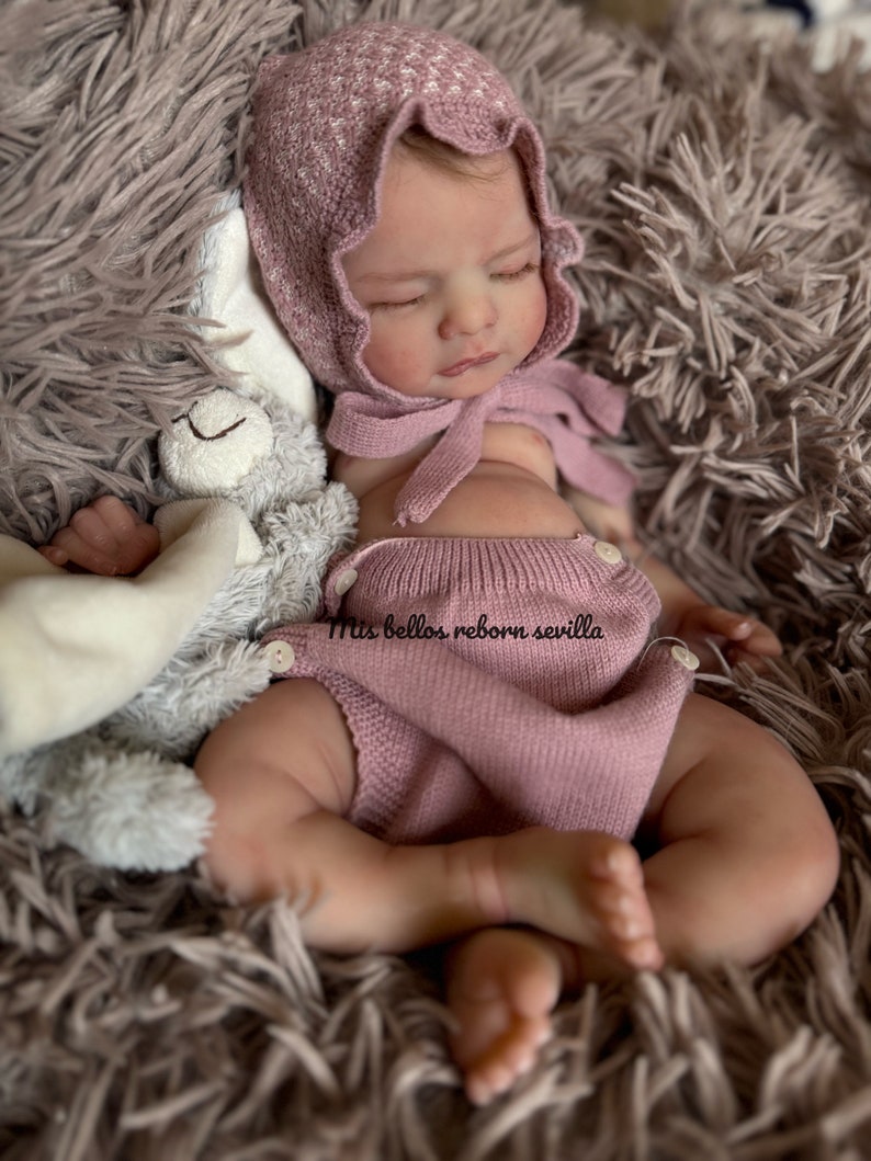 Może przedstawiać: Śpiąca lalka reborn w r&oacute;żowej dzianinowej czapce i r&oacute;żowym dzianinowym pajacyku. Lalka leży na puszystym brązowym dywanie z szarą pluszową zabawką.