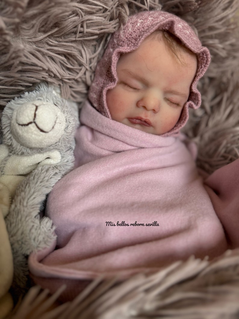 Może przedstawiać: Śpiąca lalka reborn w r&oacute;żowej dzianinowej czapce i owinięta w r&oacute;żowy kocyk. Lalka leży na puszystym szarym podłożu z szarą pluszową zabawką obok. Na kocyku widoczny jest napis "Mis bellos reborn sevilla".