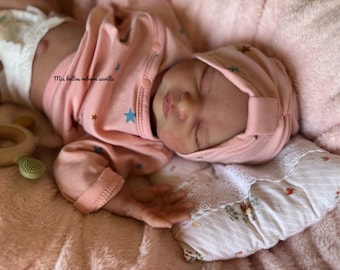 Puede incluir: Un bebé reborn durmiendo con un atuendo rosa con estampado de estrellas y un gorro rosa. El bebé está acostado en una manta rosa con una almohada blanca.