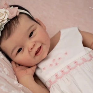 May include: A realistic baby doll with dark hair, wearing a white dress with pink floral embroidery and a matching headband featuring a cream-colored rose. The doll has a smiling expression and is branded with the text Maria Lynn Dolls.
