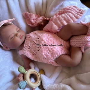 Puede incluir: Un beb&eacute; reborn durmiendo con un conjunto de punto rosa y una diadema con un lazo. El beb&eacute; est&aacute; sujetando un mordedor de madera con detalles verdes y azules. El texto "Mis bellos reborn sevilla" es visible en el conjunto del beb&eacute;.