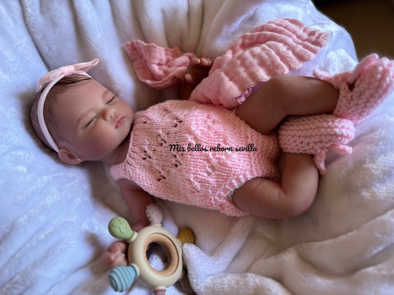 Puede incluir: Un beb&eacute; reborn durmiendo con un conjunto de punto rosa y una diadema con un lazo. El beb&eacute; est&aacute; sujetando un mordedor de madera con detalles verdes y azules. El texto "Mis bellos reborn sevilla" es visible en el conjunto del beb&eacute;.