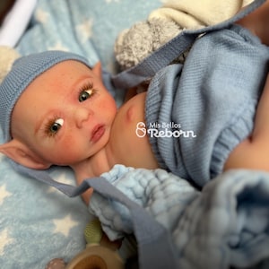 Puede incluir: Una muñeca reborn con tez clara, mejillas rosadas y pecas, que lleva un gorro de punto azul con un pompón blanco y un peto de punto azul a juego. La muñeca tiene ojos grandes y está tumbada sobre una manta azul y blanca con estampado de estrellas.